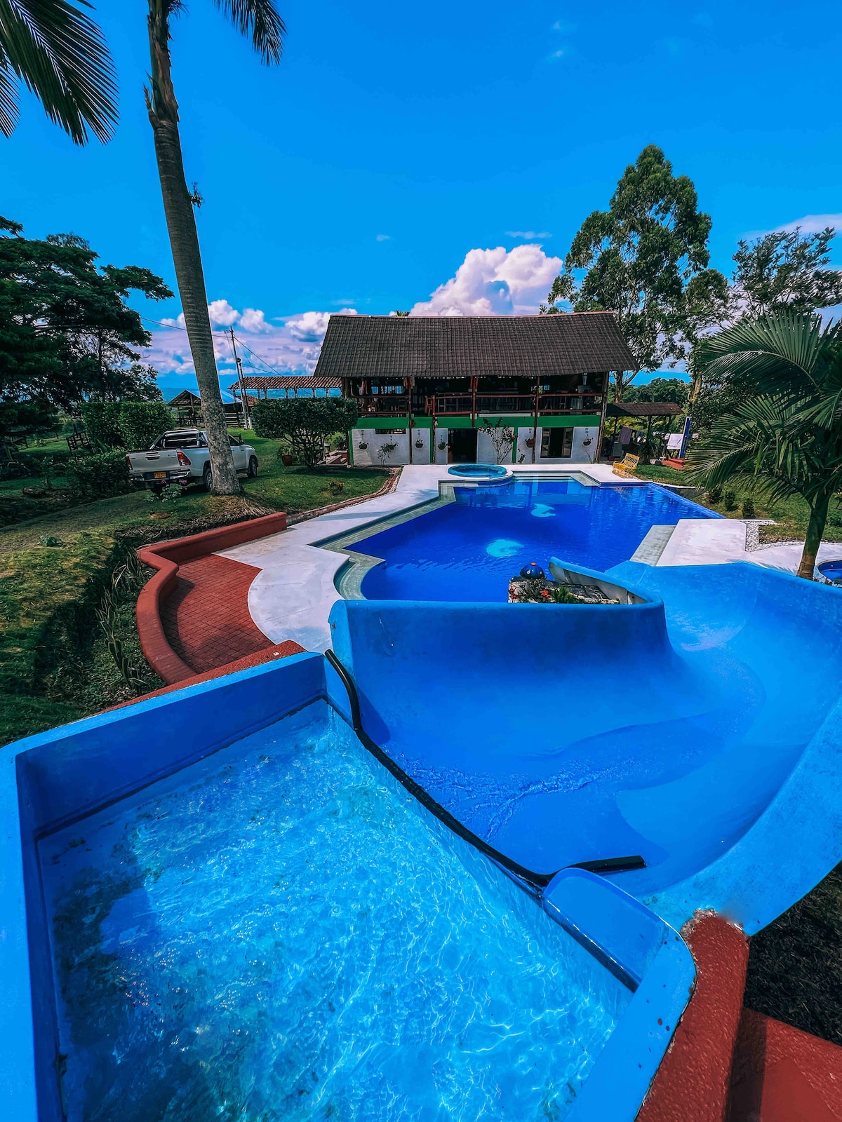 A spacious outdoor pool area features a large water slide leading into crystal-clear water. Surrounding greenery and well-maintained paths are visible, enhancing the recreational space. The inviting pool deck is complemented by a traditional-style building in the background.
