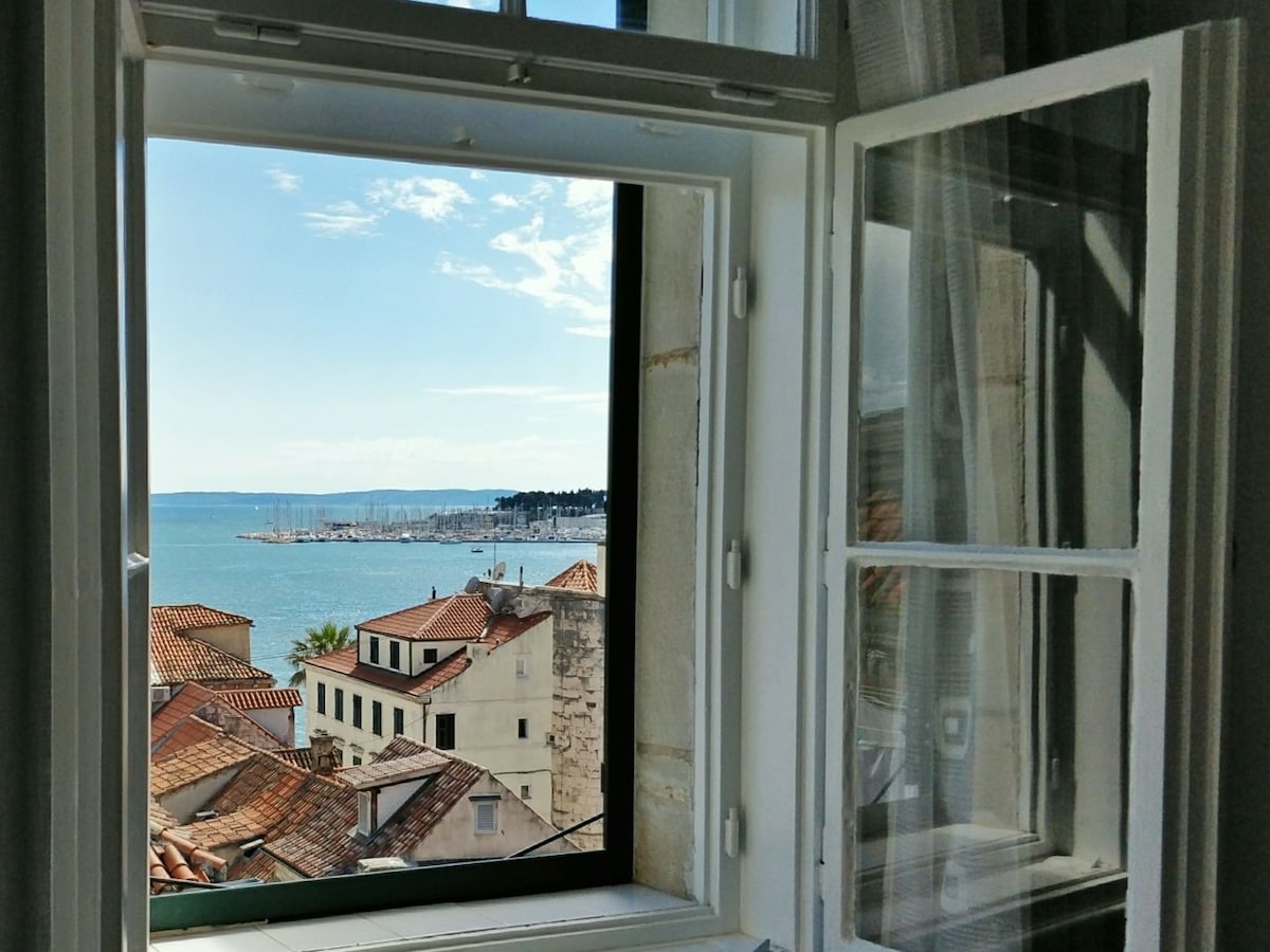 A window is framed by white shutters, revealing a stunning view of the sea. The scene includes rooftops and masts of boats in the distance, bathed in natural sunlight. The clear sky adds to the tranquil ambiance of the outdoor landscape.