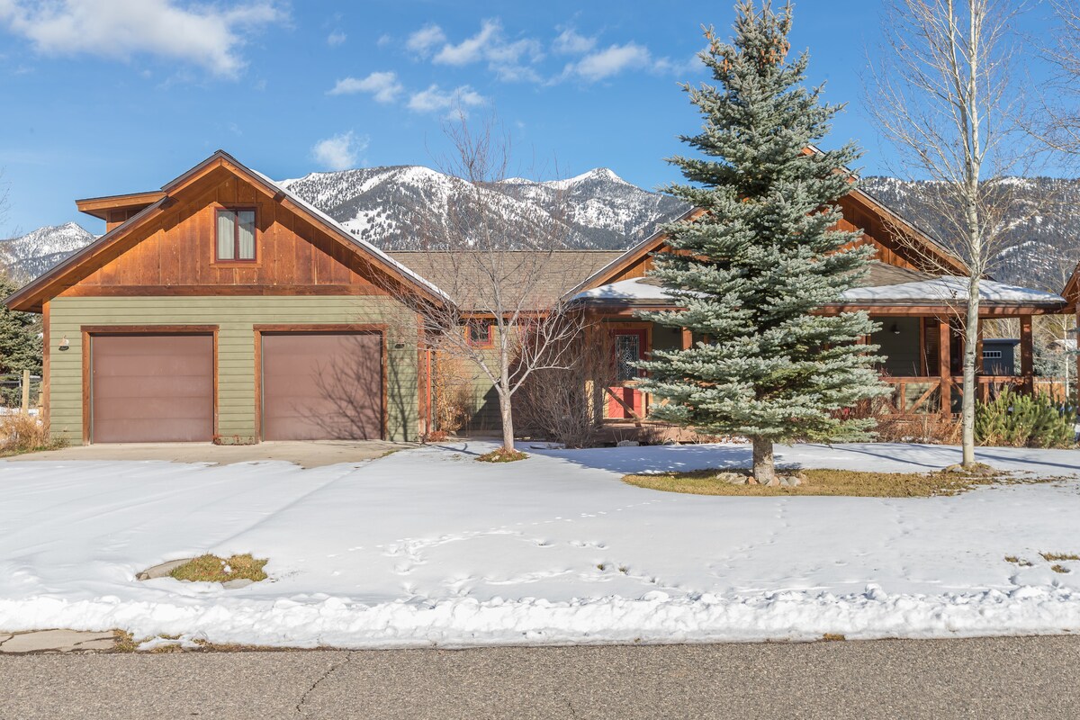 The exterior of Cold Smoke Mountain Lodge is presented, featuring a two-story wooden structure with a combination of stone and wooden siding. A snowy yard with patches of grass surrounds the home, complemented by a driveway leading to a two-car garage. Scenic mountain peaks are visible in the background.