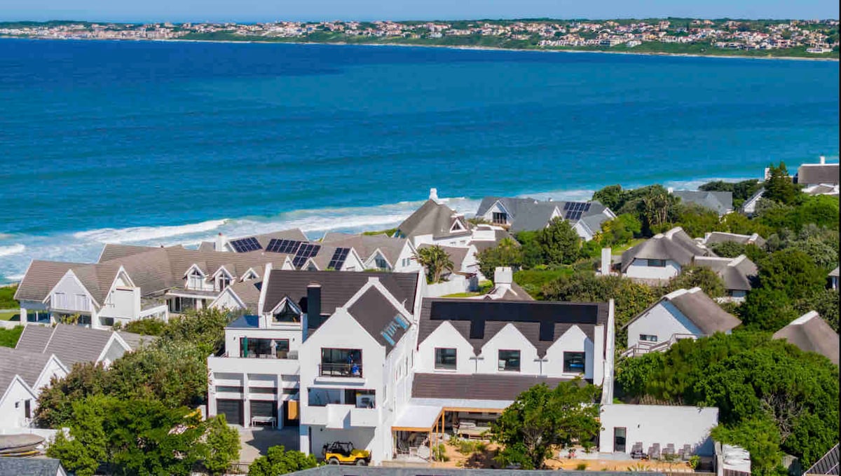 An aerial view displays a three-story white dwelling overlooking a serene ocean. The surrounding area features various rooftops, with some homes showcasing solar panels. The vibrant blue sea and sandy beach are visible, emphasizing the property’s coastal location.