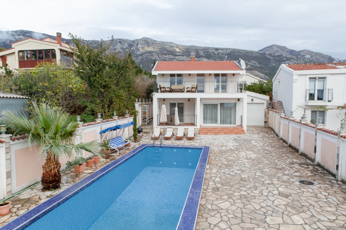 The villa is viewed from an elevated position, showcasing a large swimming pool surrounded by stone decking. Sun loungers are positioned along the poolside, with a backdrop of the two-story house and mountains in the distance. Lush greenery and potted plants enhance the outdoor setting.