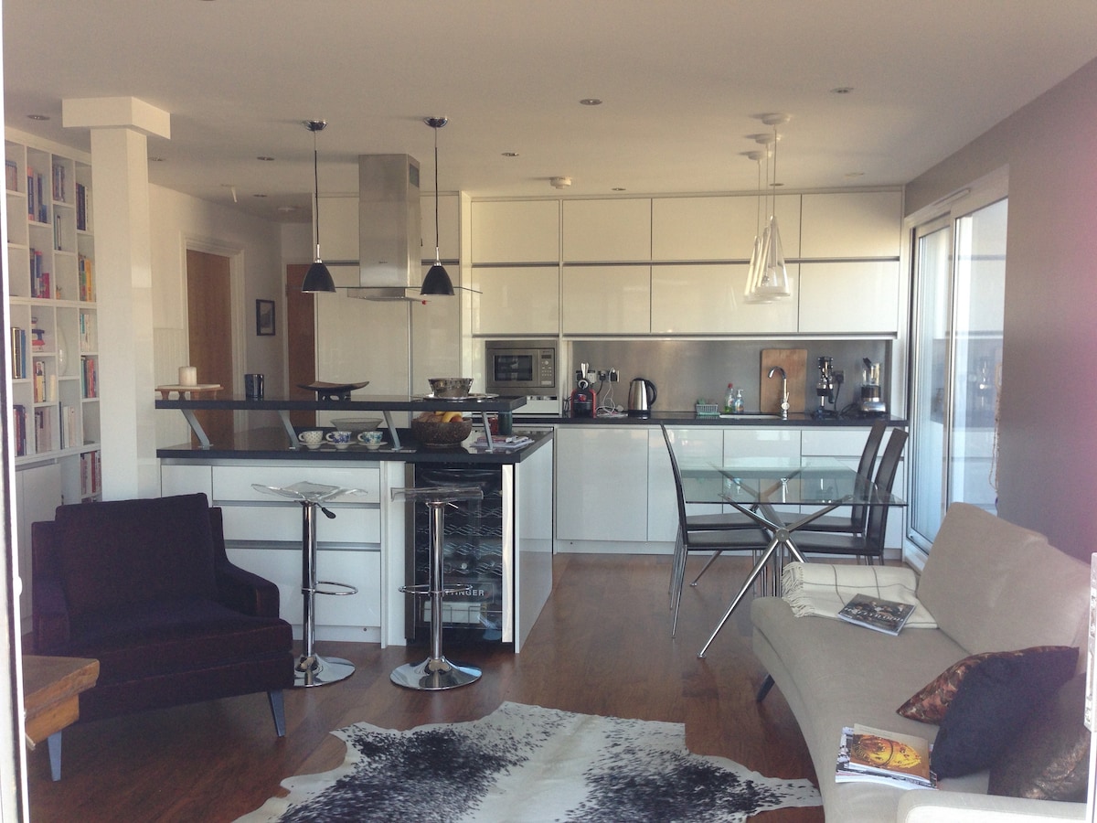 An open-plan kitchen and living area showcases sleek cabinetry and modern appliances. A glass dining table is paired with transparent chairs, while a velvet armchair and a light-toned sofa provide seating. A bookshelf lined with books adds character to the space.