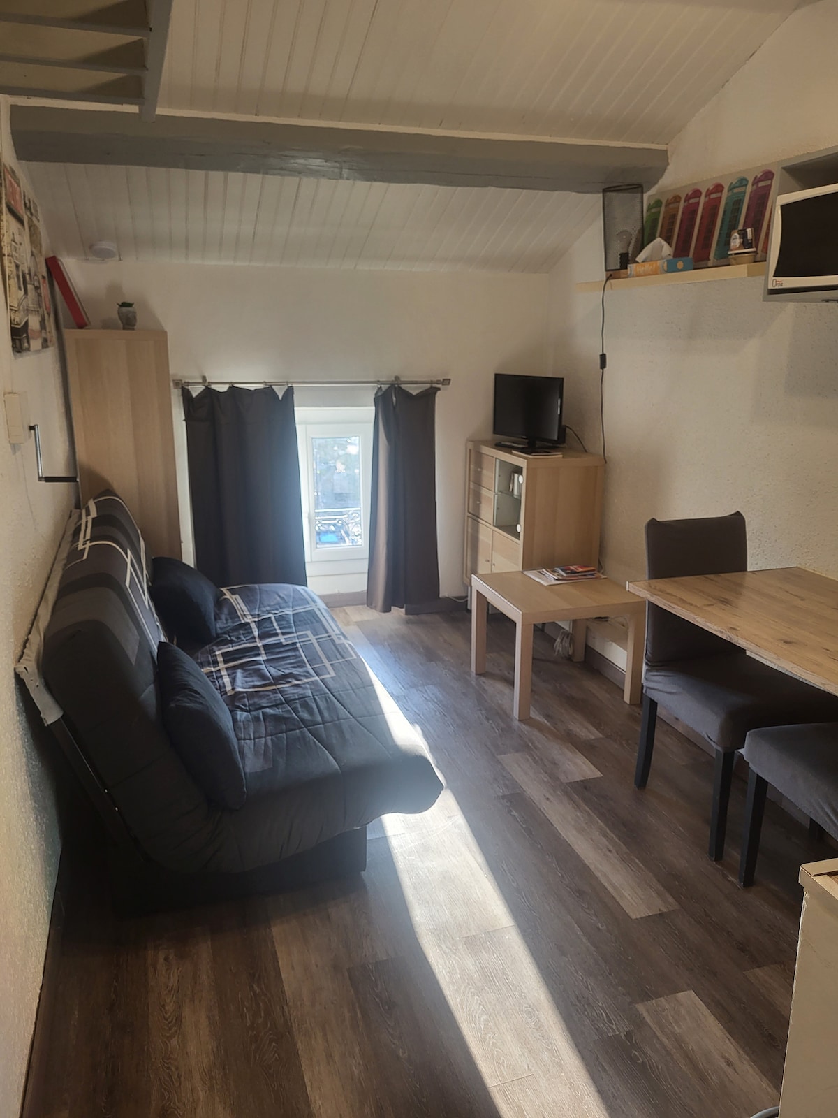 A compact living area features a sofa bed and a small dining table with two chairs. Natural light enters through a window adorned with dark curtains. An entertainment unit positioned opposite displays a television, and a kitchenette is visible in the background.