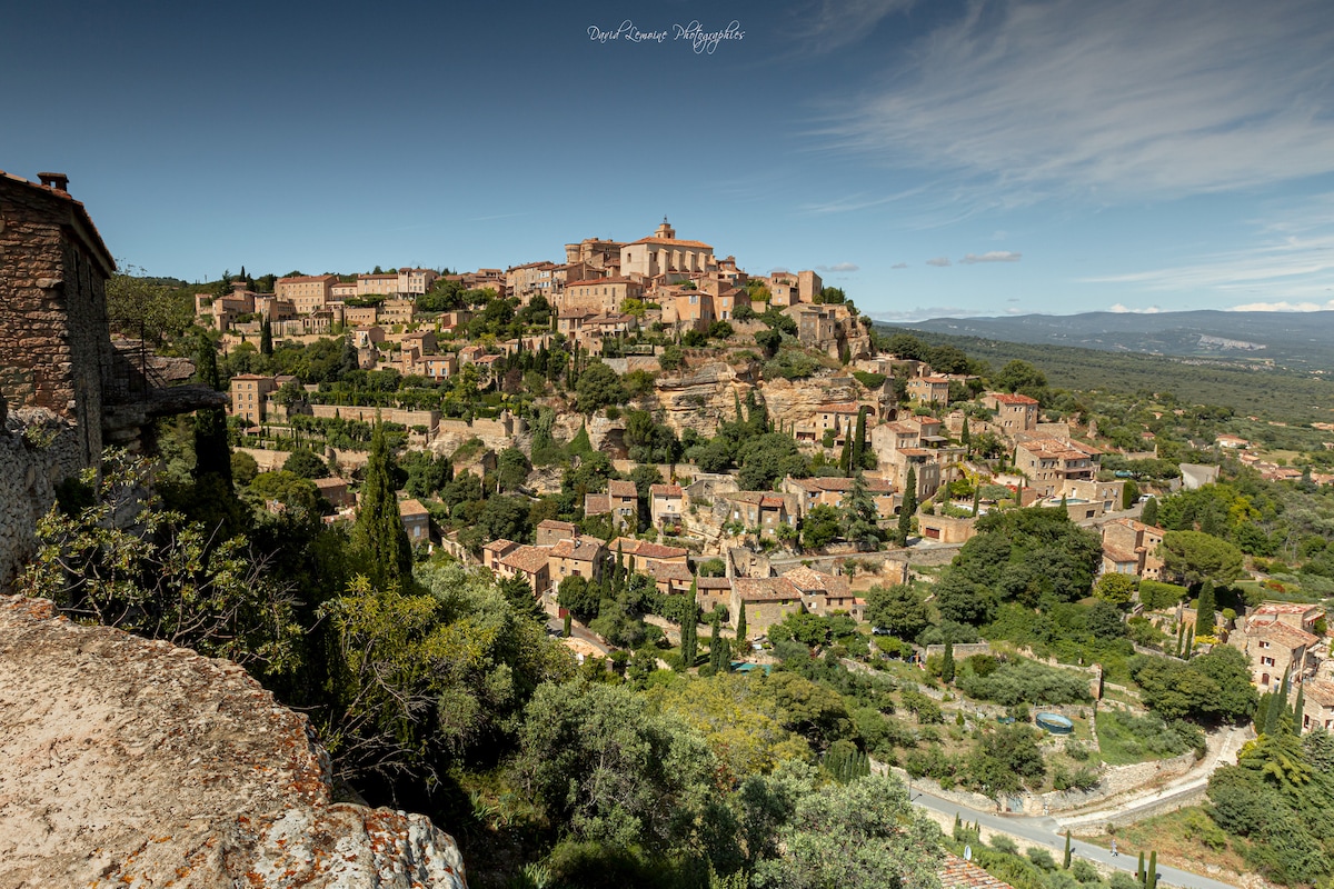 A panoramic view showcases a historic village nestled on a hilltop, surrounded by lush green landscapes and terracotta rooftops. The settlement is characterized by its natural undulations, revealing a blend of architecture among the hillside, with an expansive sky overhead.