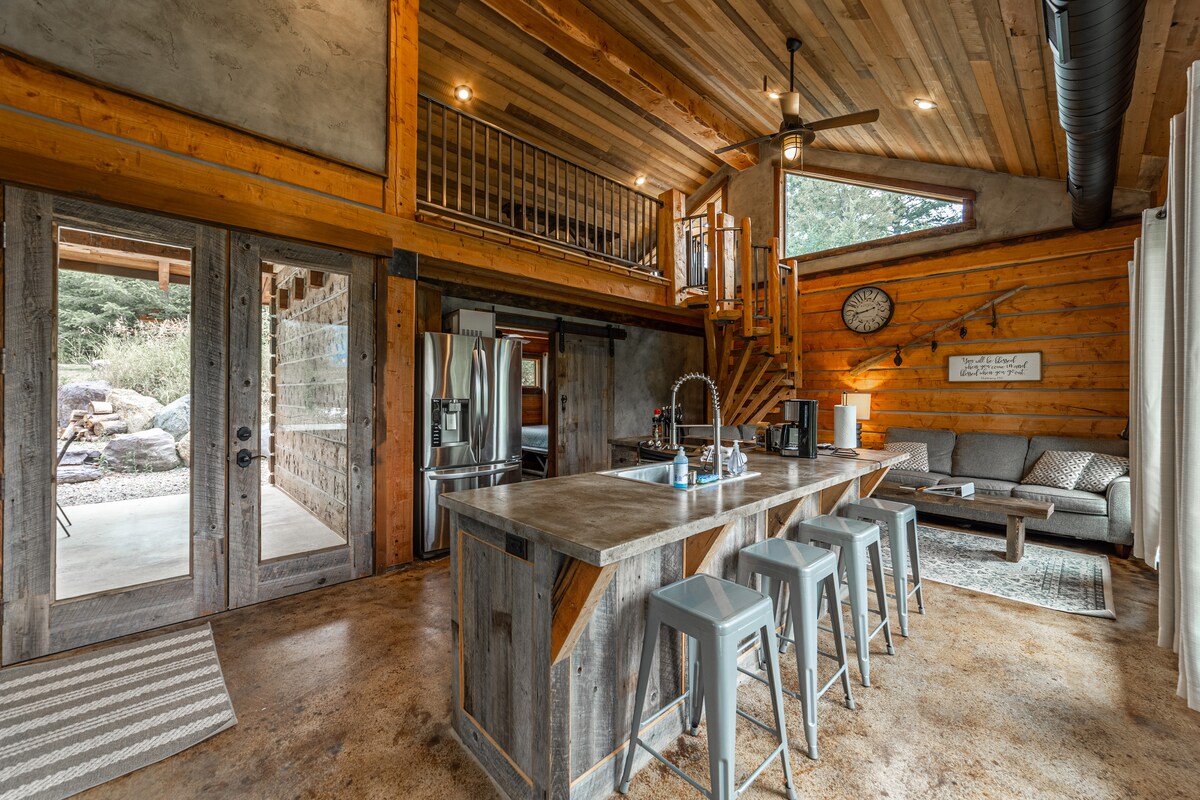 An open floor plan is presented, featuring a rustic-modern kitchen with stainless steel appliances and a large island with seating. Natural light floods the space through glass doors leading outside. The living area includes a comfortable couch and an inviting staircase to the loft above.