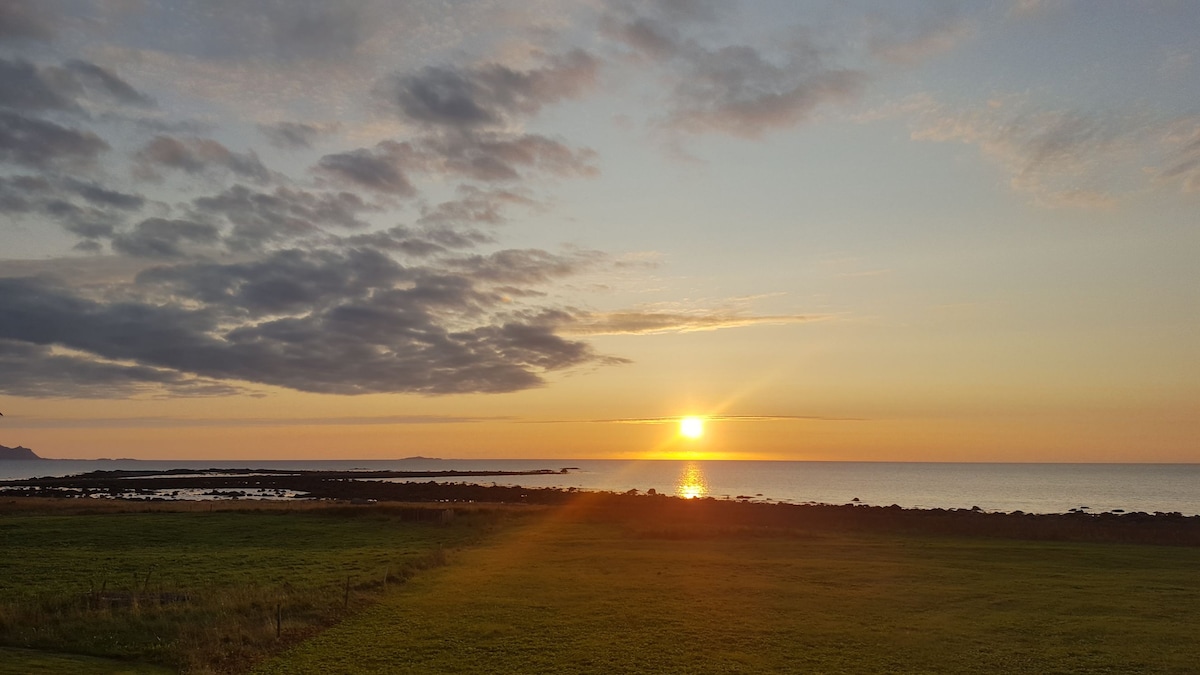 A serene coastal sunset is captured over the calm sea, with the sun dipping below the horizon. Gentle clouds are visible against a soft gradient of orange and blue, while a green expanse of grass stretches in the foreground, offering a sense of tranquility and spaciousness.