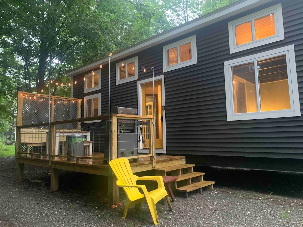 The exterior of the tiny house features a spacious wooden deck adorned with string lights, enhancing the outdoor living space. A bright yellow Adirondack chair is positioned on the deck, with multiple large windows allowing natural light to illuminate the interior.
