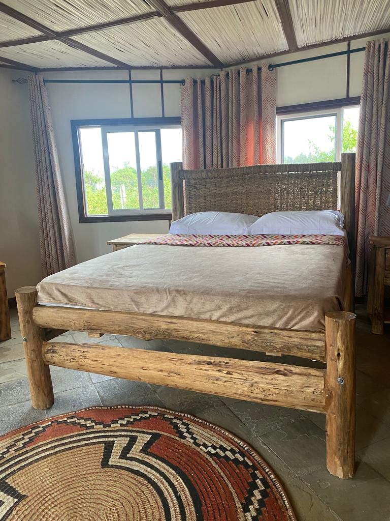 A spacious bed with a natural wood frame is centered in the room, adorned with a neutral-toned bedspread. Two large windows are positioned on one side, allowing natural light to fill the space. Wooden nightstands complement the earthy aesthetic, and patterned curtains frame the windows.