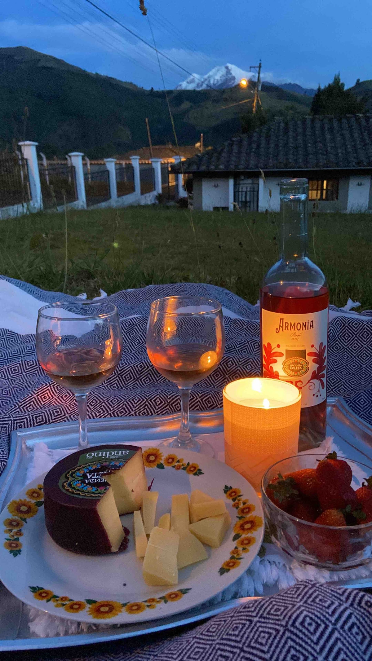 A scenic table setting features two wine glasses, a bottle of wine, and a plate with assorted cheeses and strawberries, all arranged on a patterned tablecloth. A lit candle adds a soft glow, while the snow-capped mountains serve as a tranquil backdrop.
