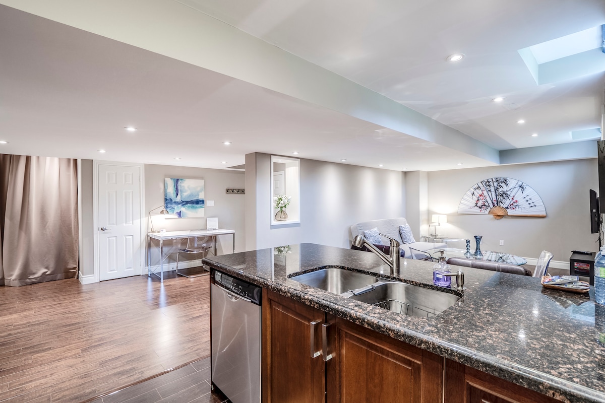 A modern kitchen features dark cabinetry and a granite countertop, with stainless-steel appliances visible. The open living area includes a comfortable sofa and a television, with large windows allowing natural light to illuminate the space. Soft colors create a calm and inviting ambiance.