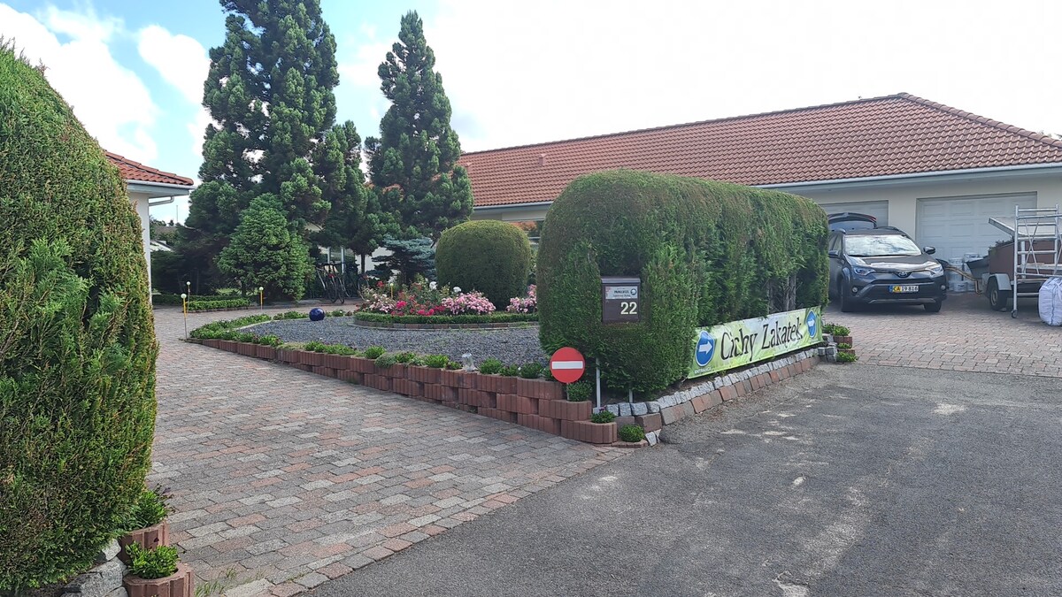 A neatly landscaped entrance features well-trimmed hedges and flower beds lining a paved driveway. A sign indicating the property name is prominently displayed, alongside a red and white no-entry sign. A spacious parking area is visible, accommodating several vehicles.