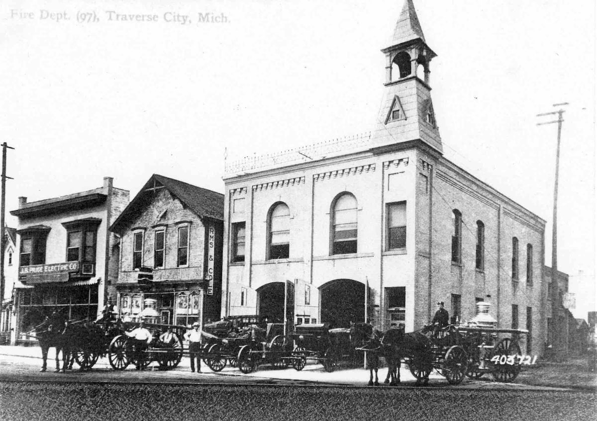 A historic black and white photo depicts the original Fire Department building in Traverse City, showcasing its iconic tower and arched windows. Horse-drawn carriages are present in front, indicating the building’s role in the community. Nearby structures capture the architectural style of the era.
