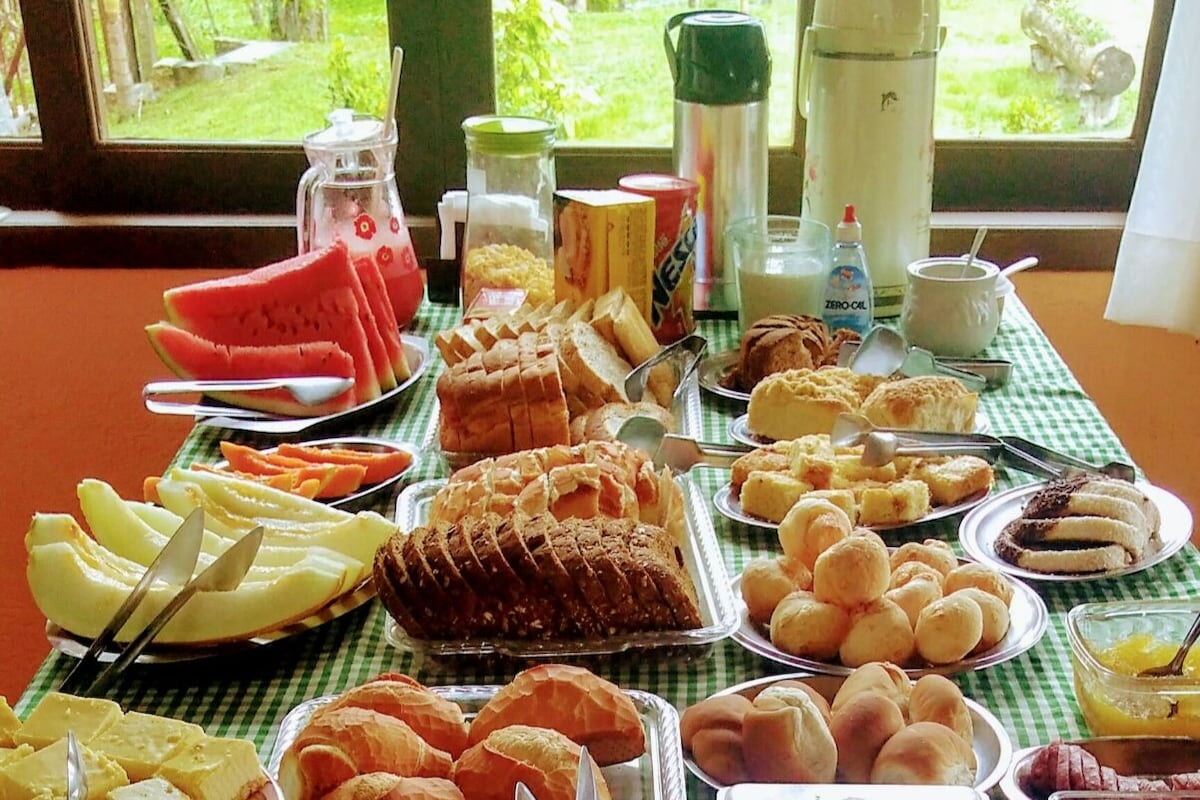 An array of fresh breakfast items is displayed on a checkered tablecloth, featuring sliced fruits like watermelon and cantaloupe. Various types of bread, pastries, and spreads are arranged neatly, along with beverages and utensils, all set against a backdrop of greenery visible through large windows.