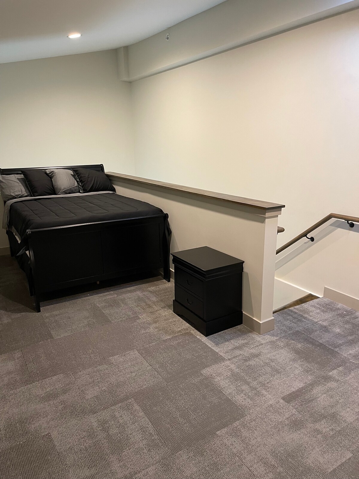 A calm sleeping area features a spacious bed with dark bedding, complemented by a pair of decorative pillows. A bedside table with two drawers provides convenient storage. Soft carpeting lines the floor, leading to a staircase with a simple railing.