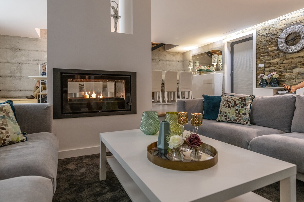 A modern living area features a stylish gray sofa arranged around a central coffee table, which holds decorative items and candles. A contemporary fireplace is positioned against a textured wall, creating a cozy focal point. Soft lighting enhances the welcoming atmosphere.