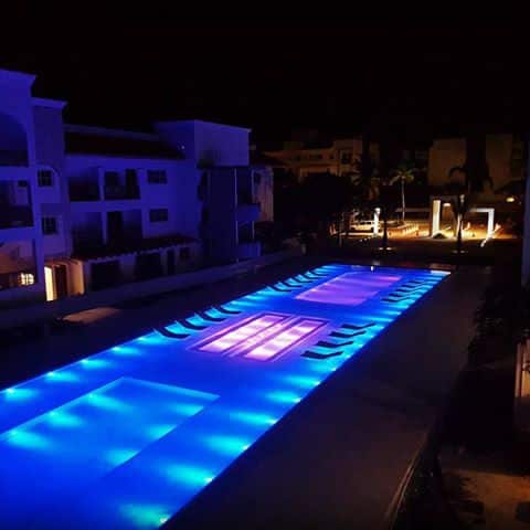 An illuminated swimming pool is showcased at night, featuring vibrant blue and purple lighting. The pool area is bordered by lounge chairs, and nearby structures are softly lit, enhancing the tranquil ambiance of the outdoor space.