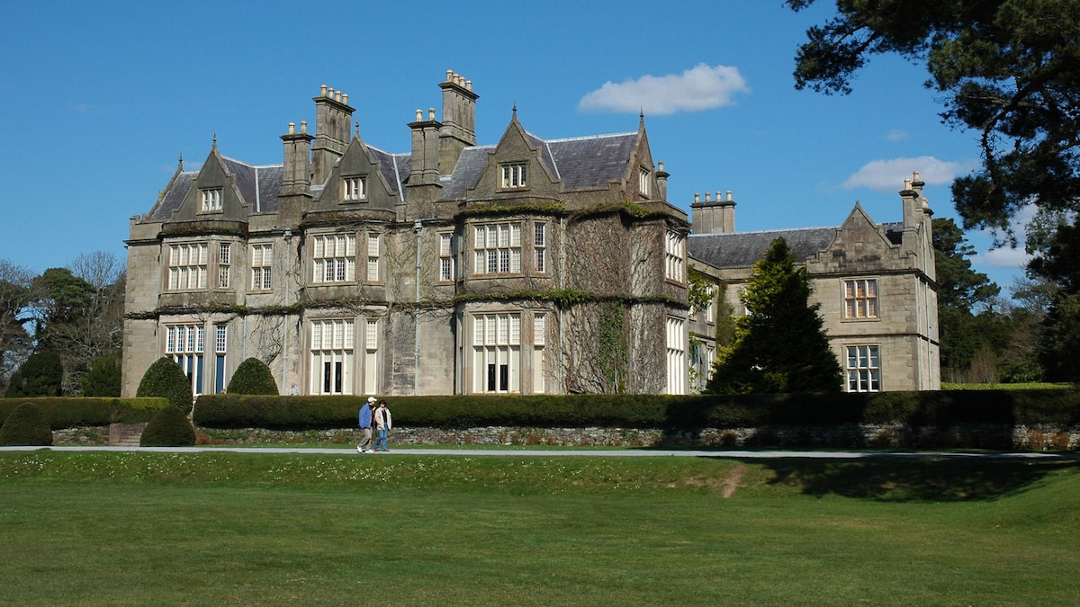 A large historic mansion is surrounded by well-maintained gardens and lawns. The building features multiple stories, numerous windows, and ornate architectural details. Tall trees and shrubs frame the property, creating a serene and expansive outdoor space.