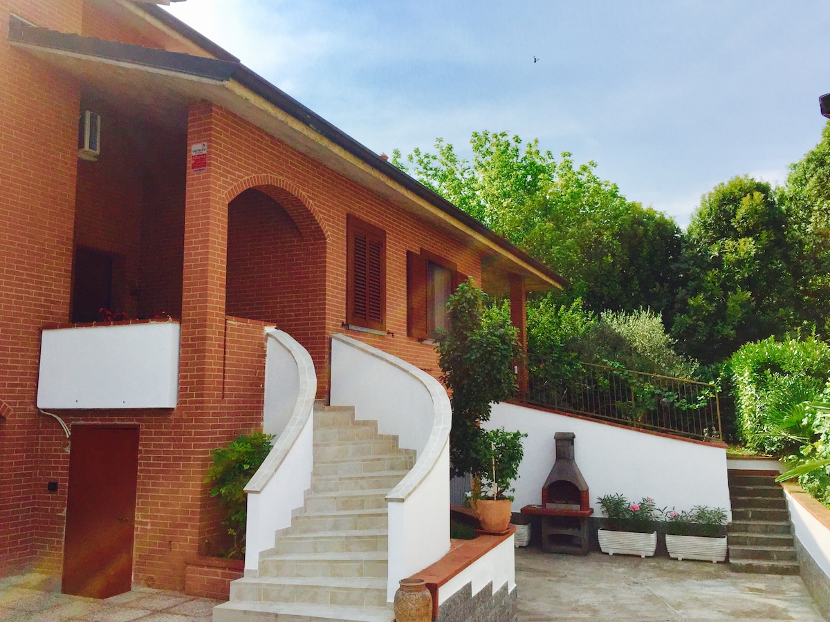 A charming villa is showcased with a large staircase leading up to the entrance. Lush greenery surrounds the property, with potted plants and a barbecue area included in the outdoor space. The warm brick facade adds an inviting touch to the exterior.