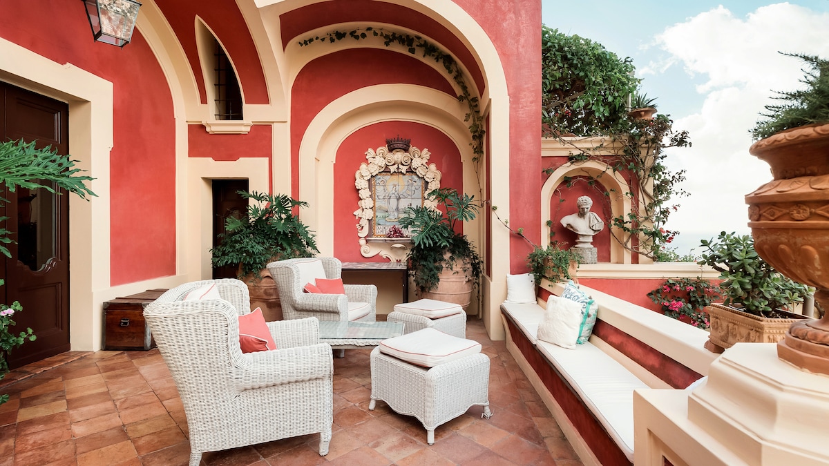 An outdoor terrace features terracotta tiles and vivid red walls, with comfortable wicker seating accented by soft pillows. Lush greenery and potted plants adorn the space, providing a serene atmosphere, while a decorative wall art piece adds an artistic touch.
