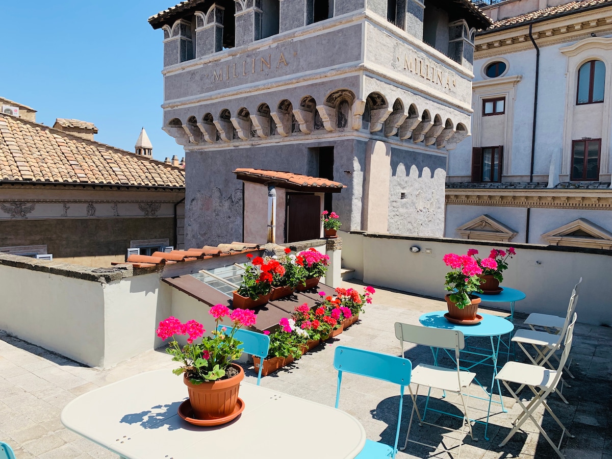 A spacious terrace is adorned with colorful flower pots, featuring vibrant geraniums. Several tables and chairs are arranged for outdoor seating, providing an inviting space to enjoy the views. The historic tower's architecture serves as a backdrop, contributing to the terrace's charm.