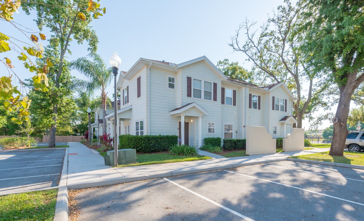 The exterior of a two-story townhouse is displayed, featuring light-colored siding and dark shutters. Lush greenery surrounds the building, with palm trees and a wide pathway leading to the entrance. Ample parking is visible in front, along with well-maintained landscaping.