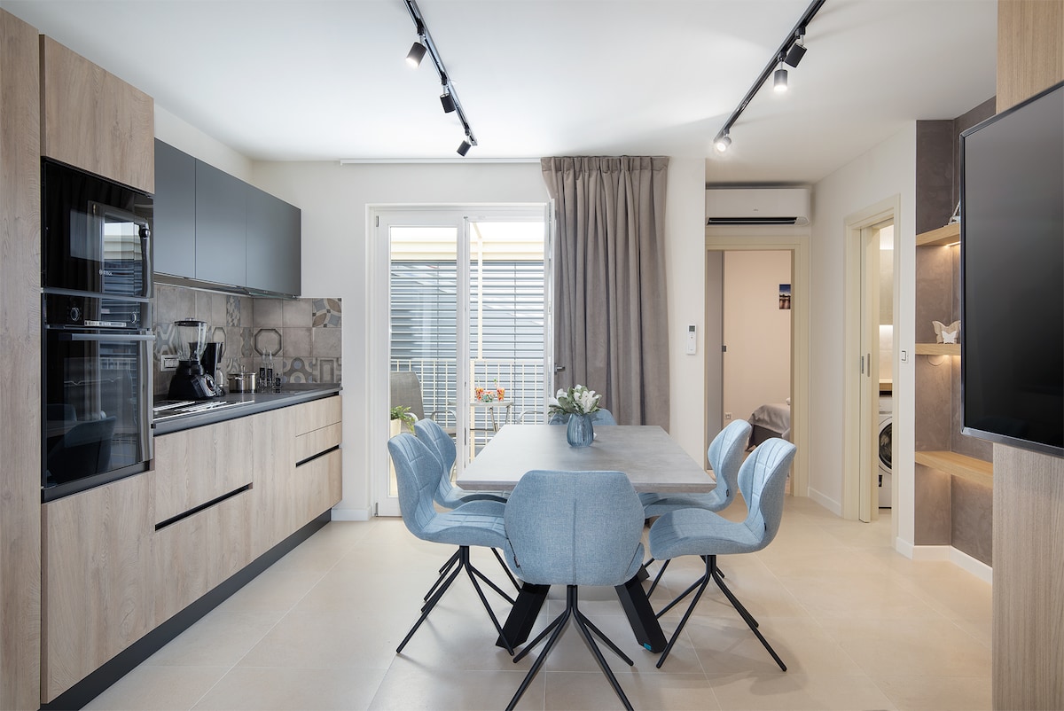 The kitchen and dining area showcases modern cabinetry and sleek stainless steel appliances. A light-colored table is surrounded by four blue chairs. Natural light filters through large glass doors leading to a balcony, with neutral-colored drapes adding a soft touch to the space.