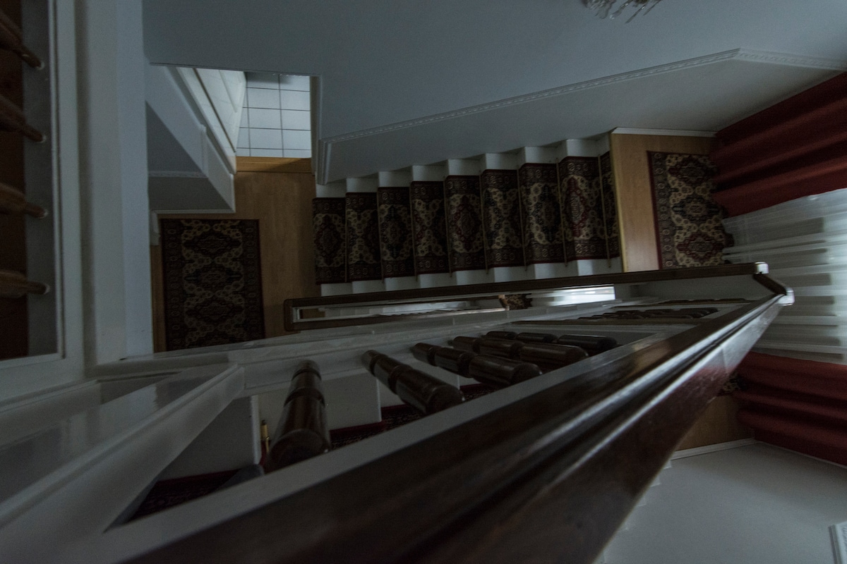 A staircase is captured from above, showcasing a wooden banister and patterned runner along the steps. The surrounding area features light-colored walls and a glimpse of a tiled floor at the top. Natural light is softened by partially drawn curtains.