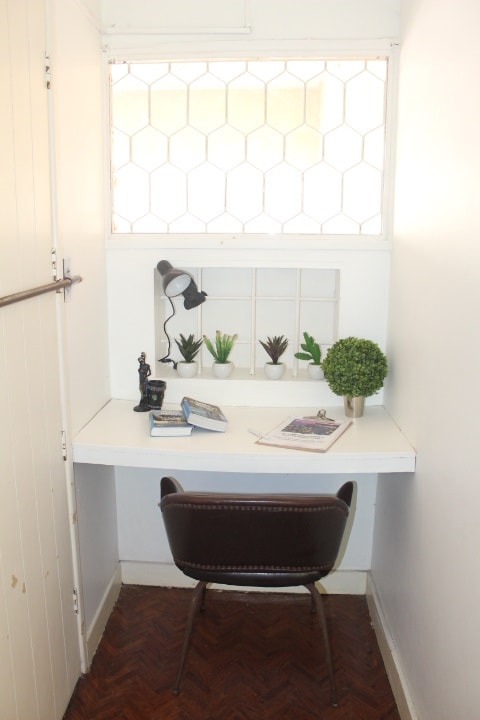 A small workspace is featured with a white desk positioned against the wall. A brown chair is placed in front of the desk, which holds decorative plants and books. A lamp provides illumination, while a window with a hexagonal pattern allows natural light to enter the space.