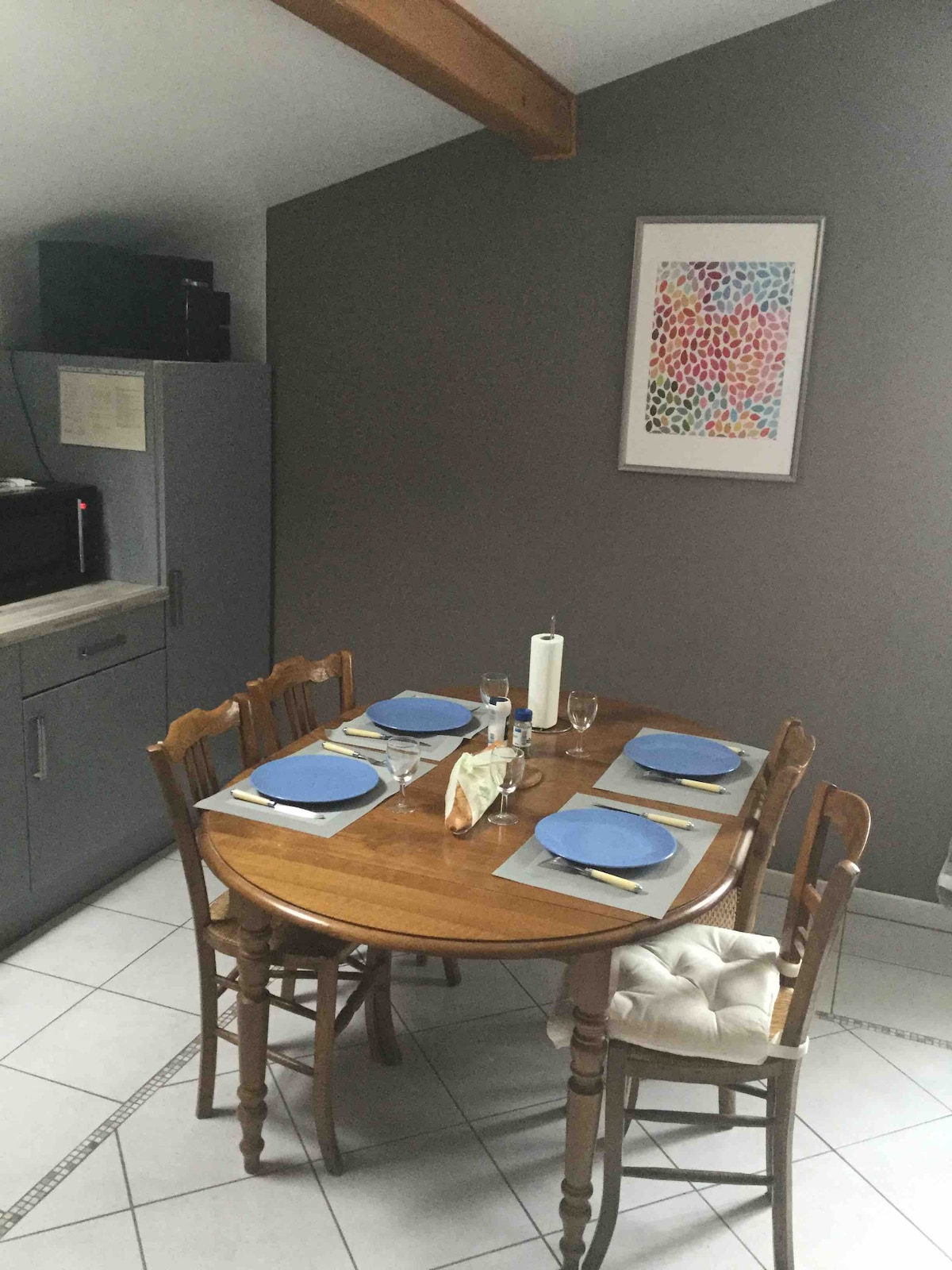 A round wooden dining table is set for four, featuring blue plates and glassware. Two wooden chairs complement the table, while a third chair has a cushioned seat. A gray wall is adorned with a colorful framed artwork, with kitchen cabinetry visible in the background.