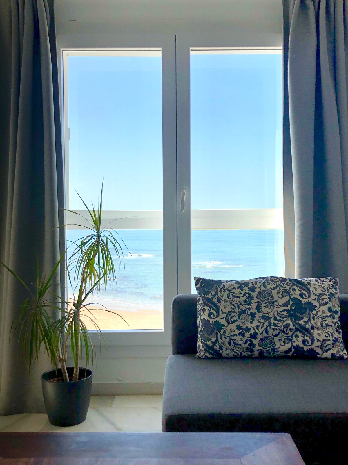 A large window offers a clear view of the beach and sea, framed by elegant gray curtains. A potted plant sits beside a cozy sofa adorned with a decorative pillow, inviting relaxation while enjoying the coastal scenery.