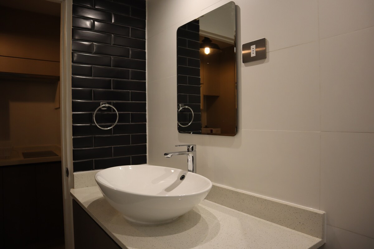 A modern bathroom features a smooth countertop with a round, white vessel sink. A rectangular mirror is mounted above the sink, reflecting the sleek black tiled wall. Minimalistic fixtures and accessories, including a towel holder, contribute to the space's clean design.