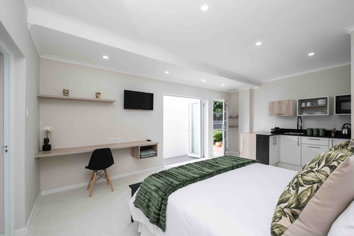 A modern bedroom area is highlighted by a spacious bed adorned with a green throw blanket. A sleek kitchenette is visible, and natural light flows in through double doors leading to an outside space. A wall-mounted television is positioned for easy viewing.