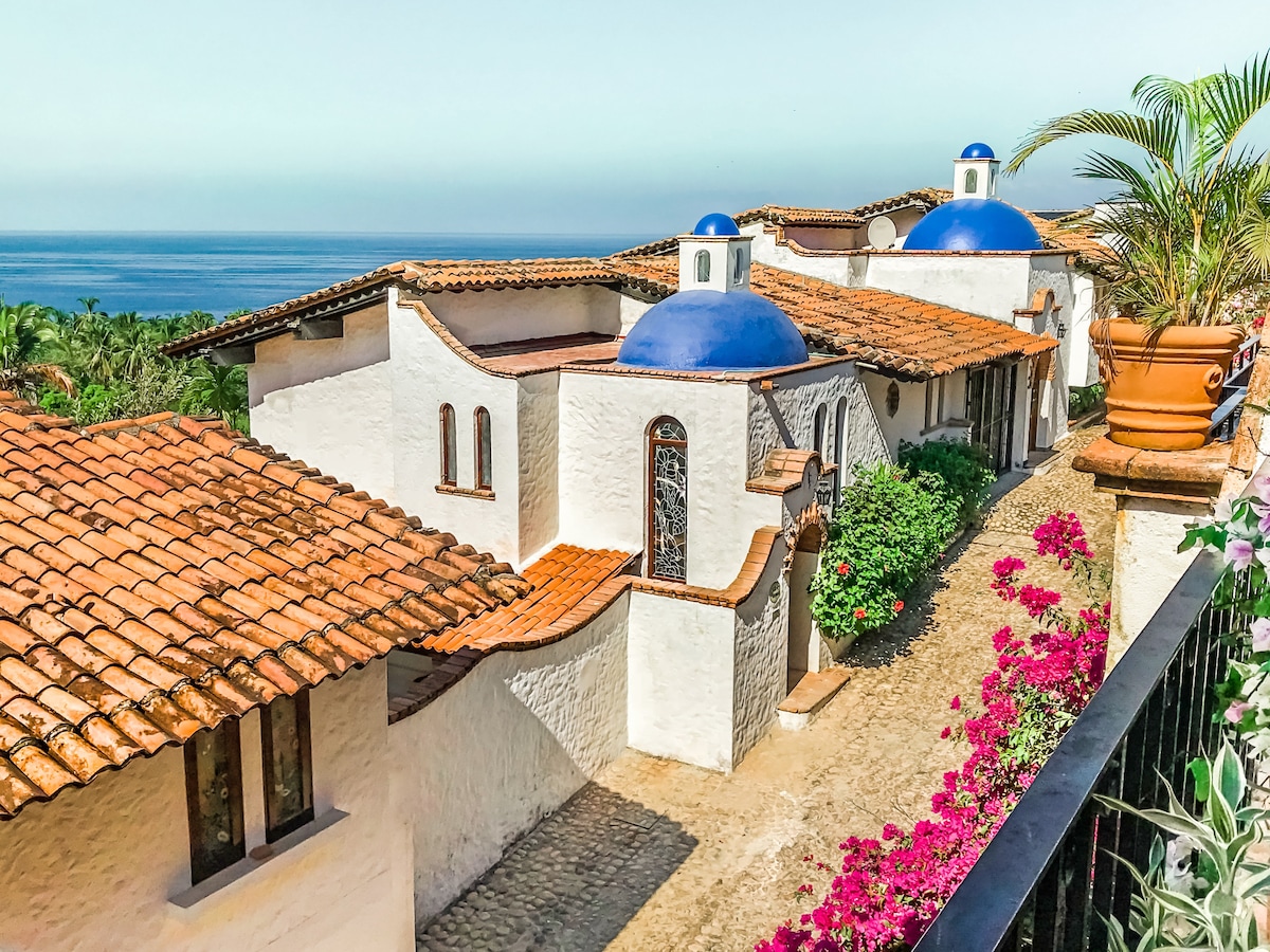 The image features a view of a Spanish Colonial villa with white stucco walls and terracotta tiled roofs. Blue domed rooftops are visible against the ocean backdrop. Vibrant bougainvillea adds a pop of color along the cobblestone walkway leading towards the ocean.
