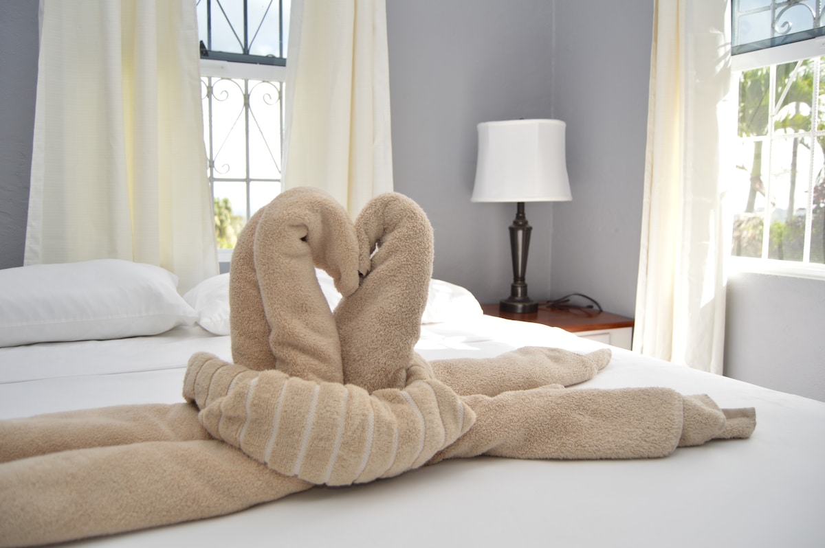 A well-made bed is adorned with two towel swans positioned gracefully in the center. Natural light enters through windows with sheer curtains, creating a soft ambiance. A bedside lamp and wooden nightstand are visible beside the bed.