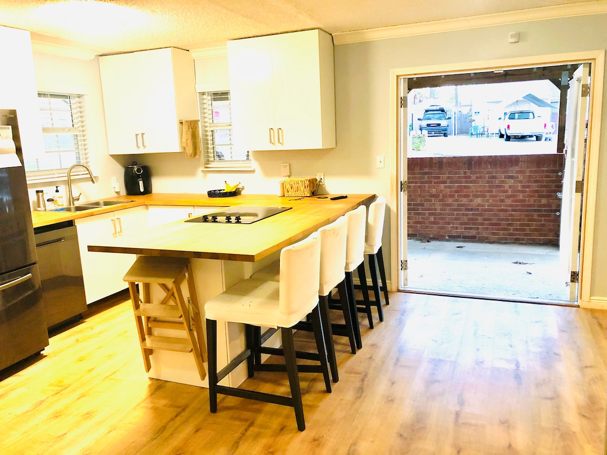 A bright open kitchen area is showcased with a central wooden island surrounded by bar stools. Light cabinets are paired with modern appliances, and a door leads to an outdoor area, allowing for easy transition between indoor and outdoor spaces.