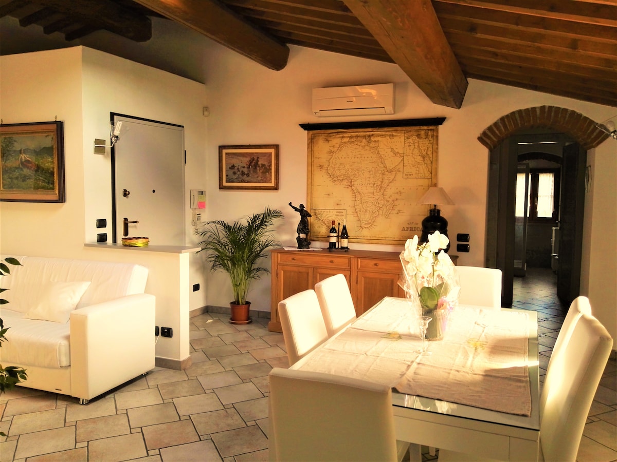 A bright living space features a white sofa, a dining table set for six, and wooden beams overhead. A decorative map is displayed on the wall, and a console table holds wine bottles and a small lamp. Potted plants add a touch of greenery.