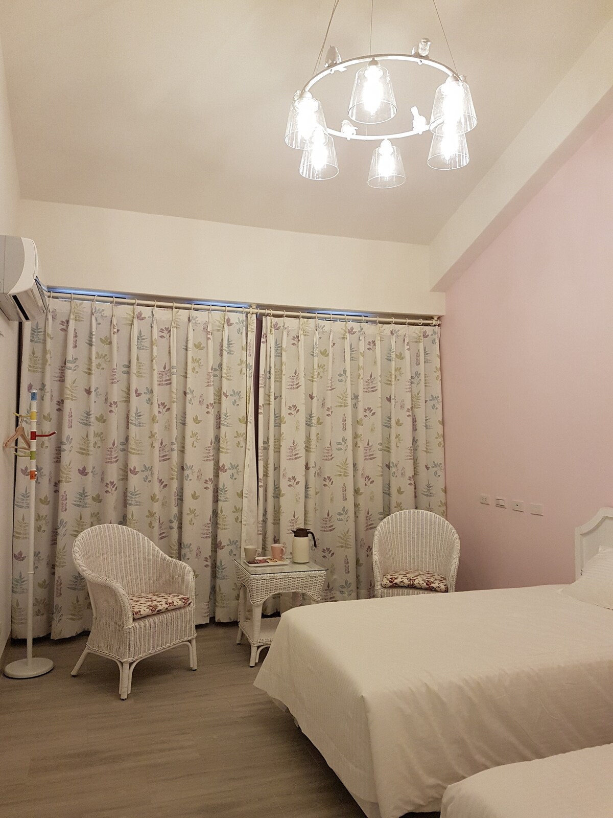 A spacious and elegantly arranged room features two beds and a set of wicker chairs adjacent to a small white table. The large window is dressed with patterned curtains, allowing for natural light while providing privacy. A modern chandelier illuminates the space.