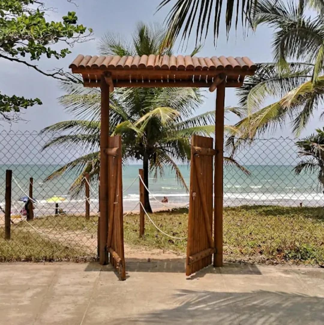 A wooden gate opens to a sandy beach, framed by lush palm trees. The ocean is visible beyond the gate, with gentle waves lapping at the shore. The area is enclosed by a fence, enhancing the sense of privacy.