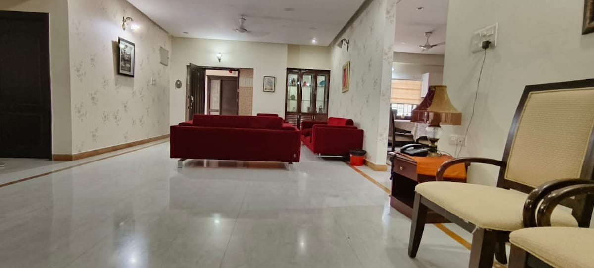 A spacious living area is furnished with red sofas arranged around a coffee table, creating a welcoming environment. Natural light is filtered through windows, enhancing the light-coloured walls and polished marble flooring. A lamp and wooden side table add functional decor to the room.
