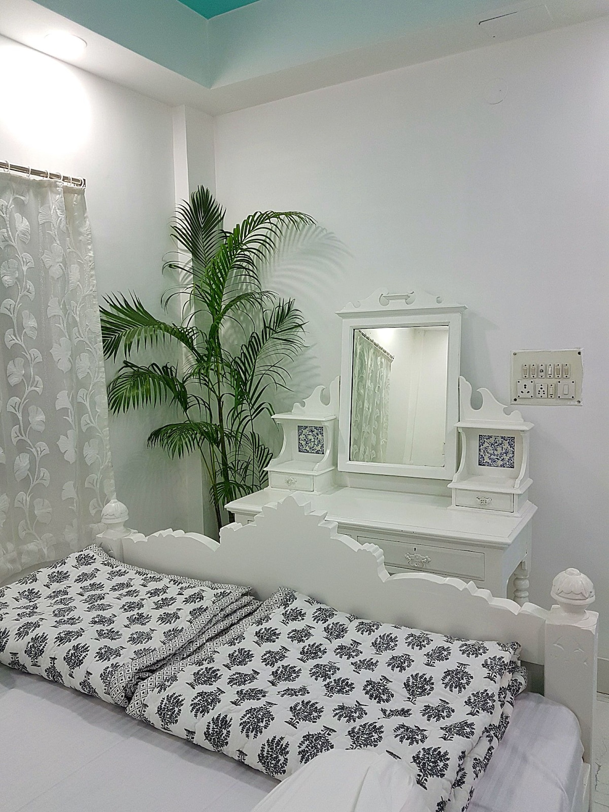 A serene sleeping area features a white wooden bed adorned with patterned pillows. A decorative mirror is positioned on a matching dresser, complemented by a small indoor palm plant. Sheer drapes softly diffuse natural light, enhancing the tranquil ambiance of the room.