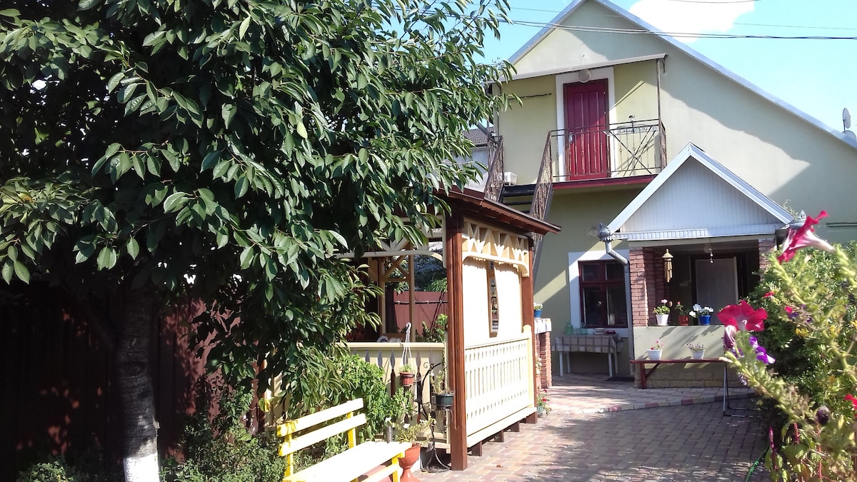 A spacious and well-maintained yard is featured, adorned with greenery and colorful flowers. A gazebo offers a cozy spot for relaxation. The house is two-storied with a balcony and exterior stairs leading to the upper floor. The separate entrance adds privacy for guests.