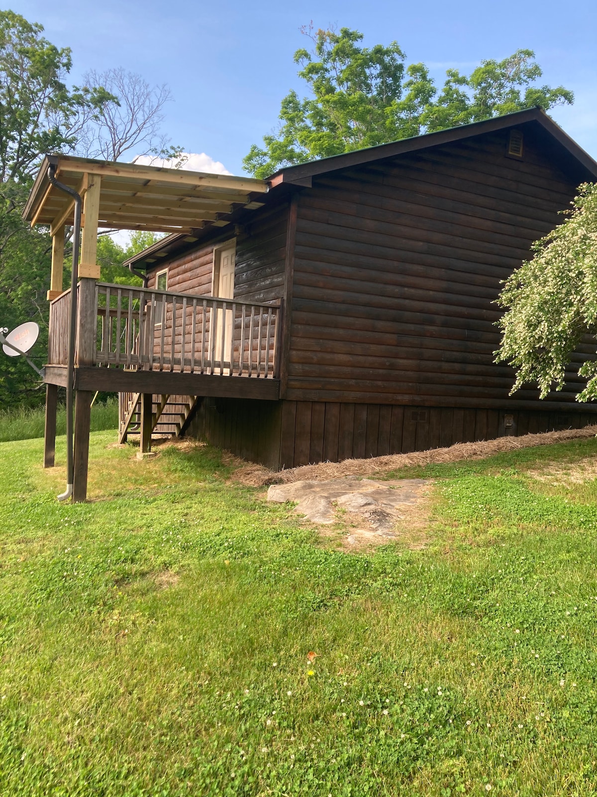 A secluded wooden cabin is set against a lush green landscape. A small porch extends from the structure, providing a shaded area. The cabin's exterior showcases a rustic log design, with natural greenery enhancing the tranquil setting.