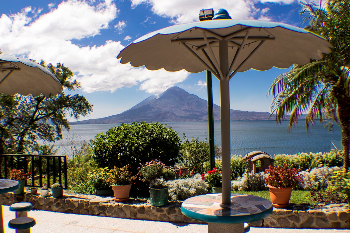 A scenic outdoor area features round tables shaded by large umbrellas. Vibrant flower pots and a variety of plants are arranged around the space, providing a colorful backdrop. A majestic volcano rises in the distance, overlooking the tranquil waters of Lake Atitlan under a partly cloudy sky.