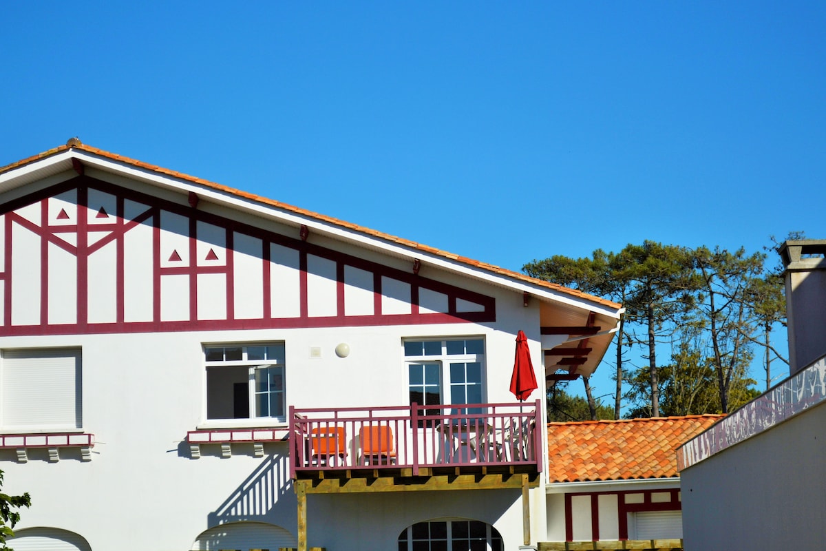 The exterior of a duplex apartment is showcased, featuring distinct structural design elements, a terracotta roof, and colorful wooden accents. A small balcony holds two chairs and a table, and a red umbrella adds a pop of color against the clear blue sky.