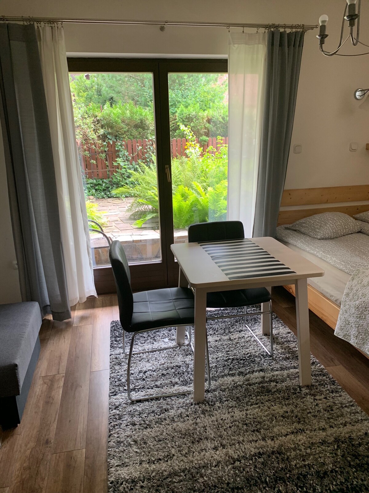 A cozy room features a bed adjacent to a dining area with a small table and two chairs. Natural light enters through a glass door that opens to a private garden, framed by sheer curtains. The space is decorated with a plush rug and warm wooden accents.