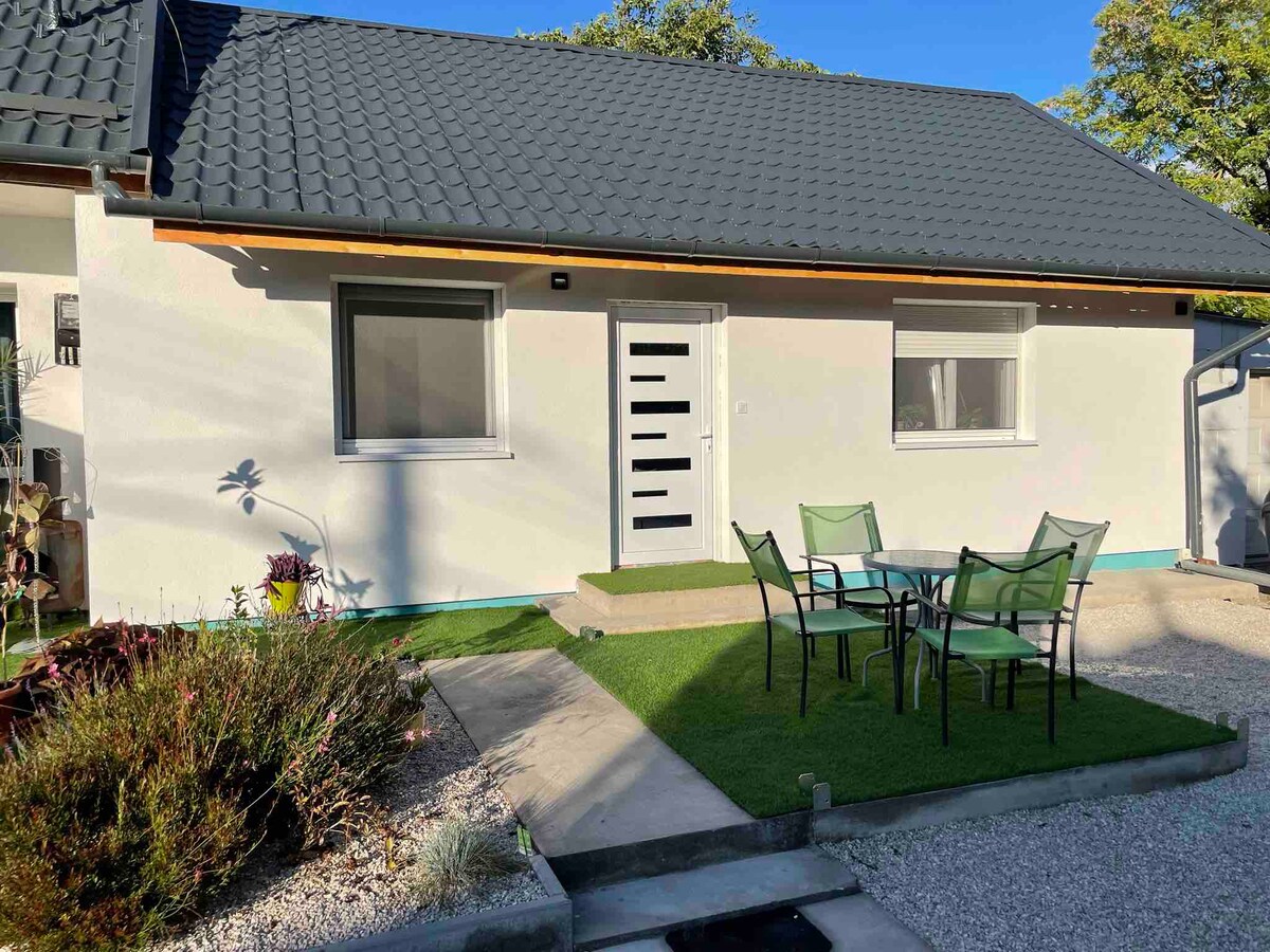 A light-colored exterior of a bungalow is highlighted by a dark, textured roof. A modern front door with a unique design provides entry, while a small area of artificial grass is bordered by a gravel path. A four-person dining table with green chairs is situated outside.