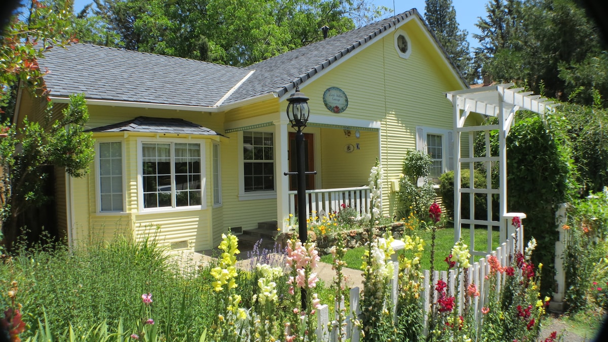 A charming yellow cottage is framed by a vibrant garden filled with blooming flowers. The front porch features railings and a decorative wall clock. Large windows invite natural light, while a trellis with climbing plants adds interest to the exterior.