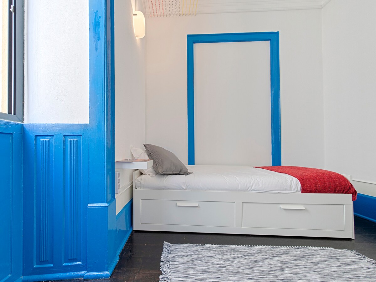 A bright sleeping area features a spacious bed topped with a neatly arranged gray and red blanket. The room is defined by bold blue accents on the walls and a white frame that draws attention to the bold color contrast.