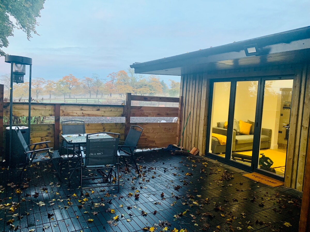 A secluded patio area is shown, featuring a four-person dining set surrounded by five chairs. The space is framed by wooden fencing, with fallen leaves scattered across the ground. Beyond the patio, a cozy lounge area is visible through large glass doors.