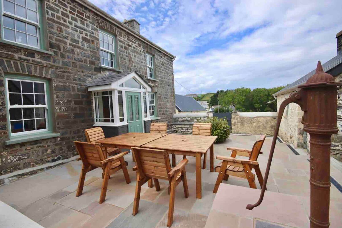 An outdoor seating area is featured, consisting of a wooden table surrounded by five matching chairs. A traditional hand pump is positioned nearby. The backdrop includes a stone building with green accents and a scenic view of the surrounding greenery.